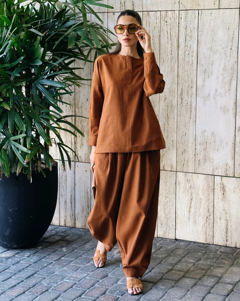 Woman in a brown outfit standing next to a potted plant against a wooden wall.