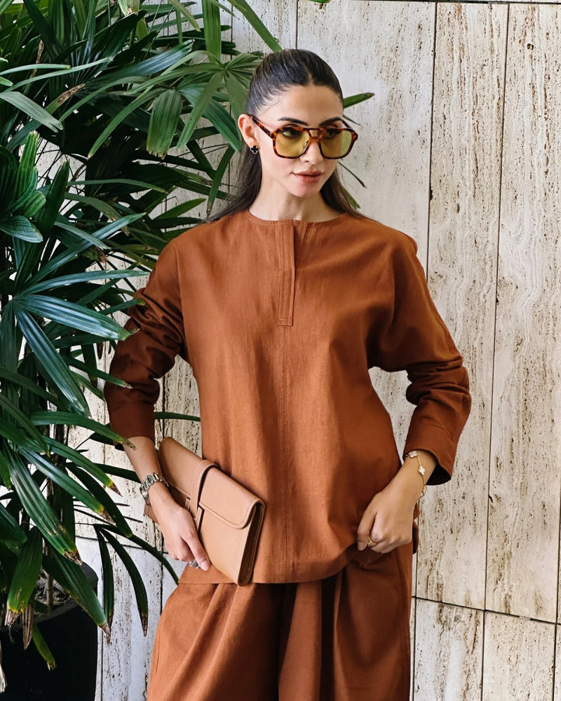 Woman in a brown outfit standing against a wooden wall with plants.