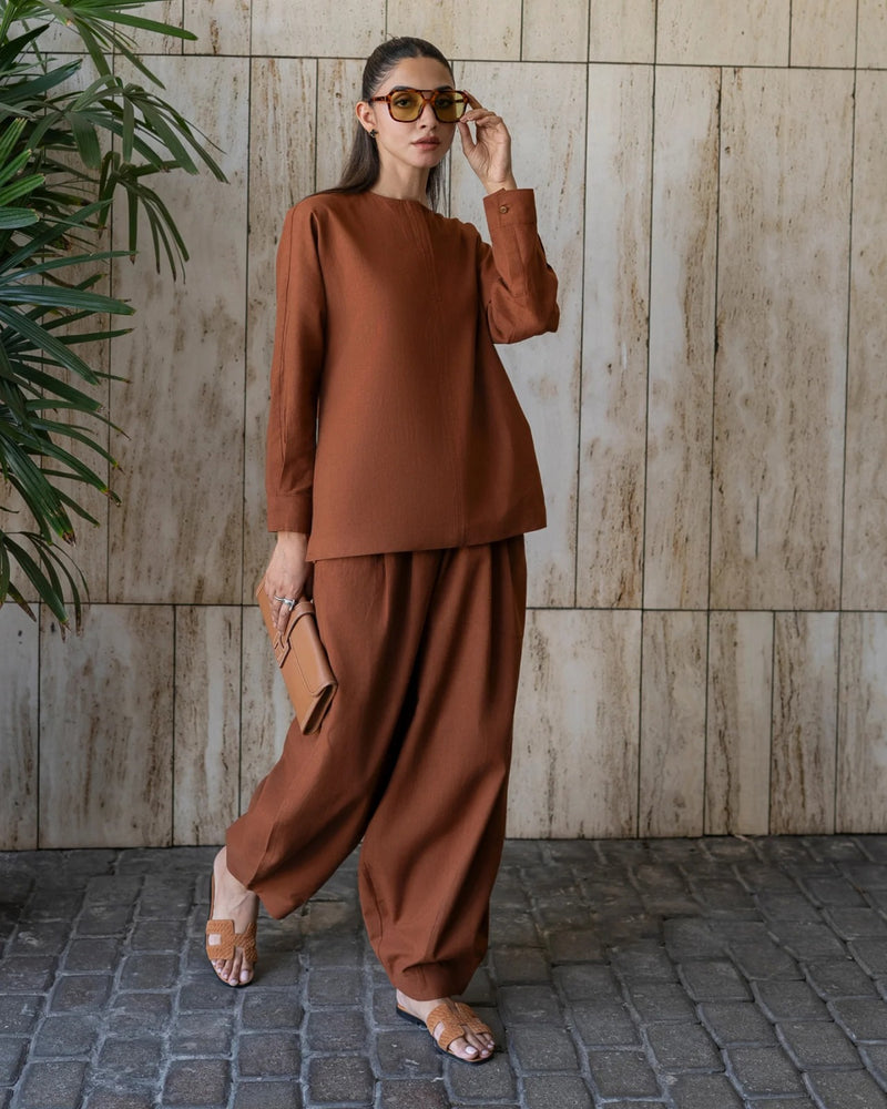 Woman in a brown outfit standing against a tiled wall with a plant to the left.