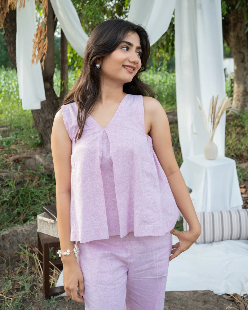 Woman in a light purple outfit standing outdoors with greenery and white fabric in the background
