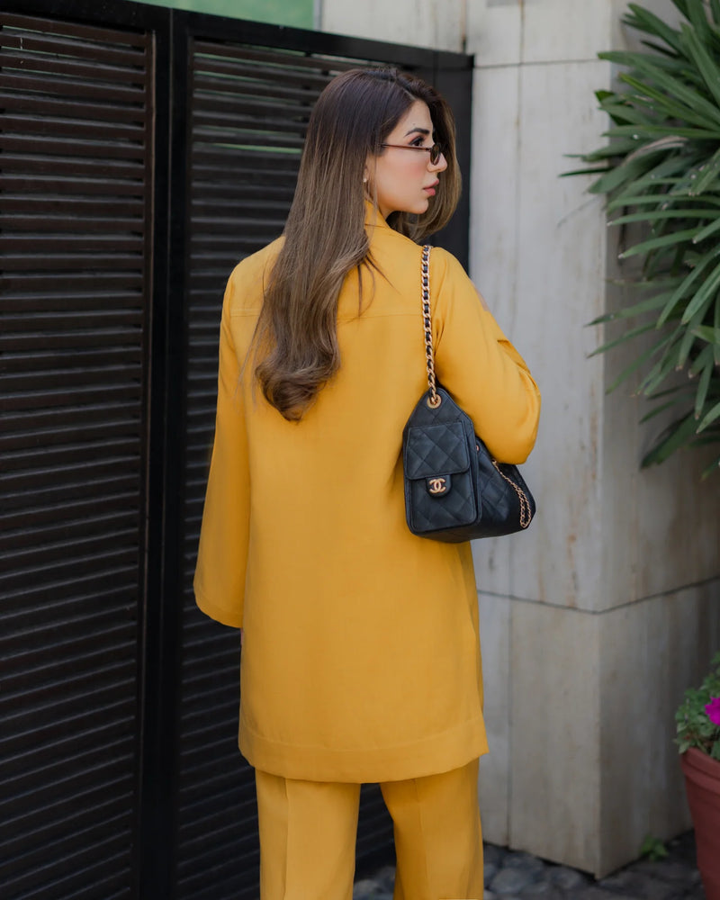 Woman in a yellow outfit holding a black handbag, standing against a building.