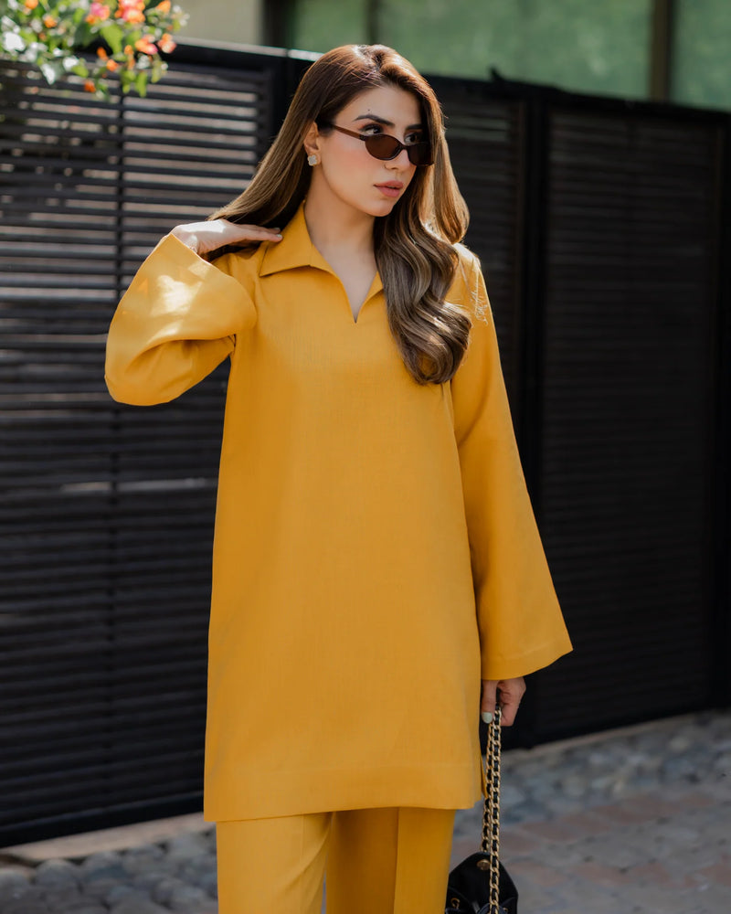 Woman wearing a yellow outfit with sunglasses, standing against a black metal fence.