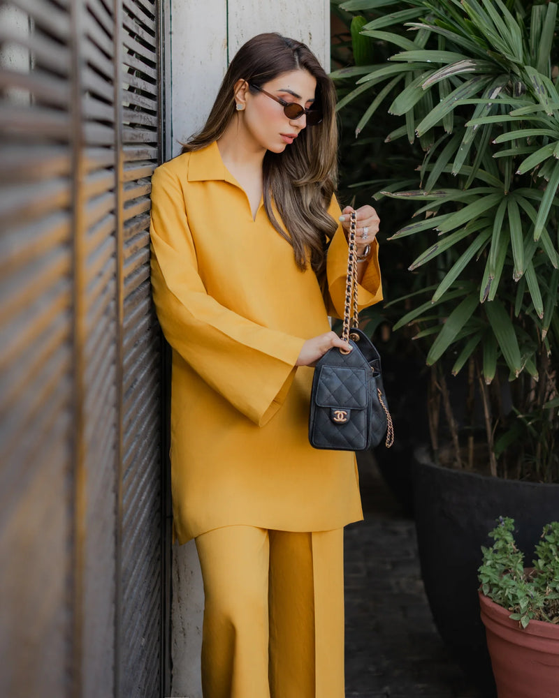 Woman in a yellow outfit holding a black handbag against a wall with plants.